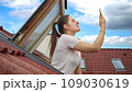 Young woman having issues with lost signal of a cellphone or smartphone looking out of the open attic window and trying to catch signal 109030619