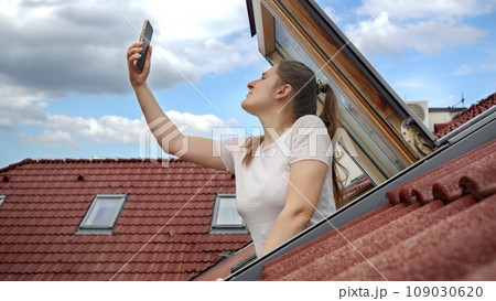 Young woman experiences signal issues with her cellphone as she peers out of an open attic window, attempting to regain a connection. Frustration of lost signal and connectivity problems 109030620