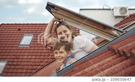 Happy smiling mother with son opens attic windows and looks outdoor. Concept of family happiness, parents with kids, moving to a new house. 109030647