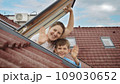 Smiling mother and her son open attic windows, gaze outside, and warmly wave towards the camera. Family happiness, parents with their children, and the excitement of moving to a new house 109030652