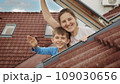 Mother and her son open attic windows, gaze outdoors, and extend a cheerful wave towards the camera. Family happiness, parents nurturing their children, and the excitement of a fresh start in a new 109030656