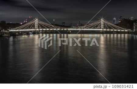 Nocturnal view of floodlit Albert Bridge from Chelsea Bridge with the reflection of the light in very calm water of River Thames. 109031421