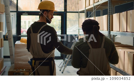 Meticulous employee walking through warehouse aisles scanning cardboard boxes on shelves under team leader guidance, using digital devices to update info before sending packages for shipping 109034852