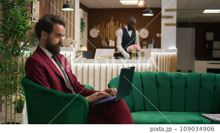 Before settling into the room, employee uses laptop in lobby reception to get organized for important presentation. Businessman heading to meeting at company headquarters while suited formally. 109034906