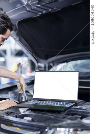 Mockup laptop on car with hood open while mechanic in blurry background replaces compressor belt. Isolated screen device next to skilled garage worker servicing client vehicle 109034949