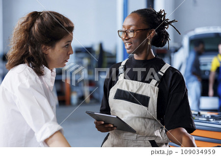 BIPOC mechanic in car service uses tablet to calculate invoice after fixing broken automobile engine. Trained auto repair shop worker uses device to inform woman of final costs after mending vehicle 109034959