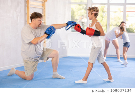 Teen boy in boxing gloves practicing punches on mitts in hands of father Teen boy in boxing gloves practicing punches on mitts in hands of father 109037134