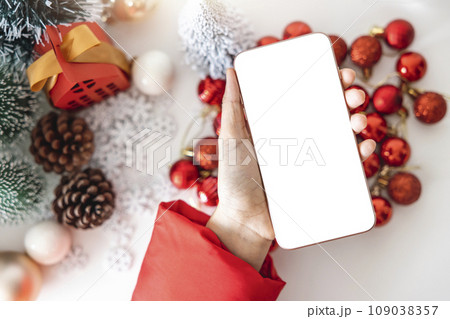 Top view Gentle hands cradle a smartphone with a white screen mock up by festive scene a beautiful Christmas tree adorned, with Christmas balls, pine cones, and red house gift box tied golden ribbon. Top view Gentle hands cradle a smartphone with a white screen mock up by festive scene a beautiful Christmas tree adorned, with Christmas balls, pine cones, and red house gift box tied golden ribbon. 109038357