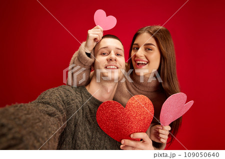 Selfie. Young happy smiling man and woman, couple, boyfriend and girlfriend posing with paper hearts against red studio background. Valentine's day, relationship 109050640