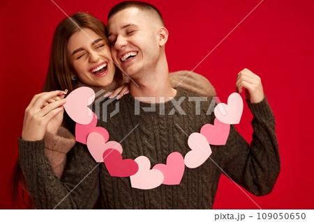 Happy, caring, loving young couple, boy and girl holding paper heart garland, celebrating holiday against red studio background. Valentine's day, relationship 109050650
