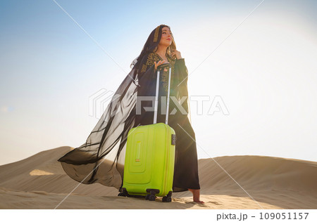 Arabian woman in traditional abaya costume with bright light green or yellow suitcase in sands. Concept and idea of travel to United Arab Emirates, sand dunes in desert of Dubai at sunset. 109051157