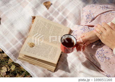 A young beautiful woman in a dress and a round hat reads a book outdoors in the forest and drinks tea. Romantic and vintage photo of a beautiful girl. Reading and relaxation 109051160