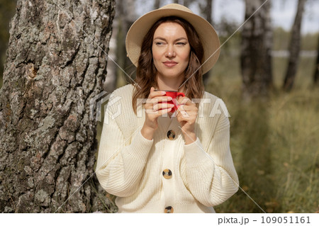 A young beautiful woman in a dress and a round hat reads a book outdoors in the forest and drinks tea. Romantic and vintage photo of a beautiful girl. Reading and relaxation 109051161