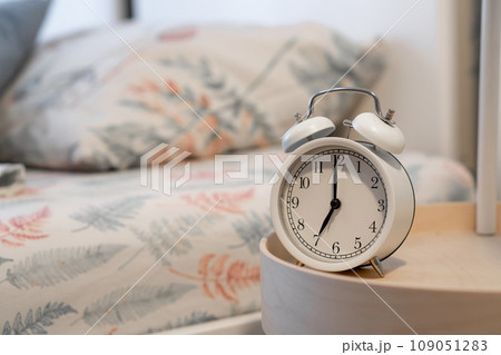 Close-up of a round white alarm clock on a table in the bedroom. The hands on the clock show seven o'clock in the morning, time to get up. Retro alarm clock on the table, vintage tone. space for text Close-up of a round white alarm clock on a table in the bedroom. The hands on the clock show seven o'clock in the morning, time to get up. Retro alarm clock on the table, vintage tone. space for text 109051283