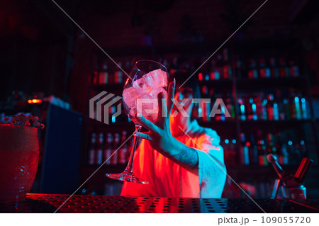 Glass with ice cubes standing on bar table. Neon colored bar space. Preparation of Aperol Spritz cocktail Glass with ice cubes standing on bar table. Neon colored bar space. Preparation of Aperol Spritz cocktail 109055720