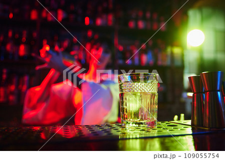 Focus on glass with ice cube. Young woman, bartender making delicious sweet and sour gin garden cocktail in bar in neon light 109055754