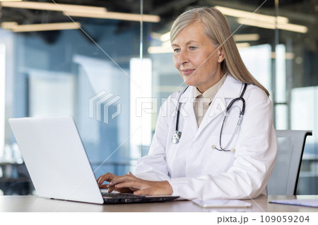 Senior gray haired female doctor in white coat and stethoscope sitting and working in clinic office at laptop online. Senior gray haired female doctor in white coat and stethoscope sitting and working in clinic office at laptop online. 109059204