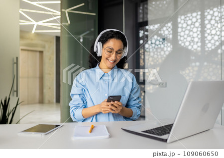 Young beautiful woman at workplace joyful listening to music and online radio, using app on phone, businesswoman resting on break, singing along well done job well done. Young beautiful woman at workplace joyful listening to music and online radio, using app on phone, businesswoman resting on break, singing along well done job well done. 109060650