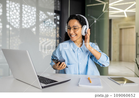 Young beautiful woman at workplace joyful listening to music and online radio, using app on phone, businesswoman resting on break, singing along well done job well done. Young beautiful woman at workplace joyful listening to music and online radio, using app on phone, businesswoman resting on break, singing along well done job well done. 109060652