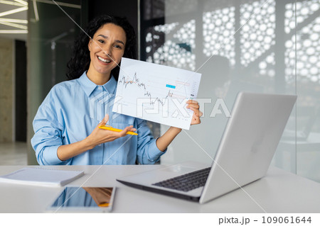 Portrait of a young successful female financier at the workplace inside the office, the businesswoman shows the graph with positive results of the enterprise's achievement to the camera. 109061644