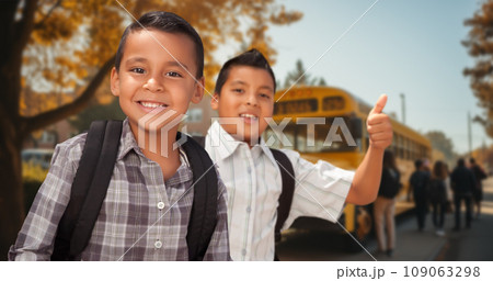 Two Happy Young Hispanic Boys Wearing Backpacks Give a Thumbs Up on Campus Near a School Bus 109063298