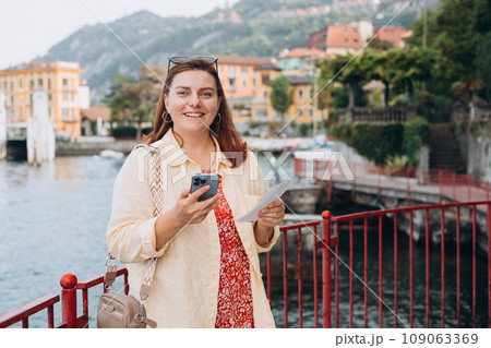 Hipster girl with two tickets browsing Internet. Cruise ship vacation concept. Varenna, Italy. Lake Como. Phone Communication. Female holding phone and booking hotel 109063369