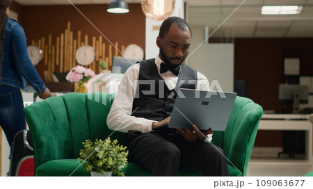 Focused hotel manager working on laptop sitting in lobby, looking at screen and keeping track of reservations to manage bookings. Bellboy in uniform works in hospitality industry. Handheld shot. 109063677