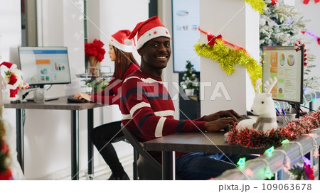 Portrait of friendly african american startup employee working on christmas season in decorated office. Workers solving tasks at workstation in multiethnic workplace during winter festive time Portrait of friendly african american startup employee working on christmas season in decorated office. Workers solving tasks at workstation in multiethnic workplace during winter festive time 109063678