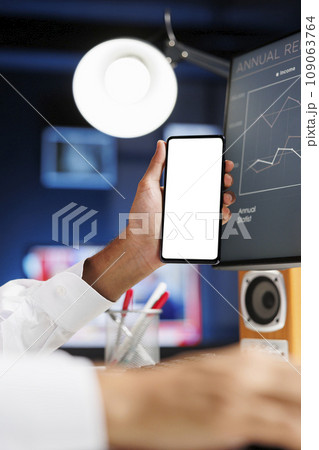 Detailed view of a male hand holding a mobile device with an isolated copyspace white screen in front of a desktop pc. Arab person displaying a blank chromakey mockup template on a smartphone. Detailed view of a male hand holding a mobile device with an isolated copyspace white screen in front of a desktop pc. Arab person displaying a blank chromakey mockup template on a smartphone. 109063764