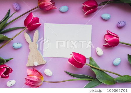 White empty envelope copy space Easter eggs and wooden bunnies with pink tulips on violet pastel background. Greeting card Postcard. Flat lay composition top view. Decoration minimalist modern design 109065701