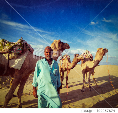 Vintage retro hipster style travel image of Rajasthan ndian man cameleer (camel driver) portrait with camels in dunes of Thar desert with grunge texture overlaid. Jaisalmer, Rajasthan, India 109067874