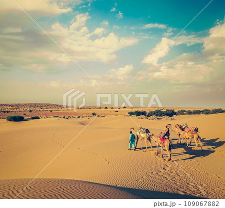 Vintage retro hipster style travel image of Rajasthan travel background - two Indian cameleers (camel drivers) with camels in dunes of Thar desert. Jaisalmer, Rajasthan, India Vintage retro hipster style travel image of Rajasthan travel background - two Indian cameleers (camel drivers) with camels in dunes of Thar desert. Jaisalmer, Rajasthan, India 109067882