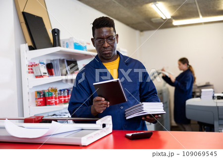 Portrait of friendly male employee of printing house with stack of notebooks in her hands Portrait of friendly male employee of printing house with stack of notebooks in her hands 109069545