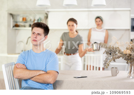 Upset guy sitting in kitchen on background of dissatisfied wife and mother-in-law Upset guy sitting in kitchen on background of dissatisfied wife and mother-in-law 109069593