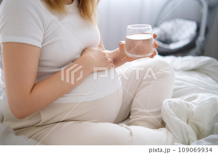 Pregnant woman sits on the bed, tenderly embracing her growing belly and holding glass of water. Pregnant woman sits on the bed, tenderly embracing her growing belly and holding glass of water. 109069934