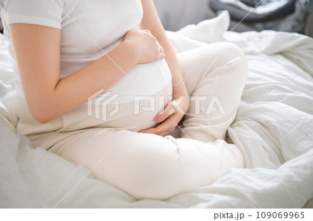 Pregnant woman takes a seat on the bed, tenderly embracing her growing belly 109069965