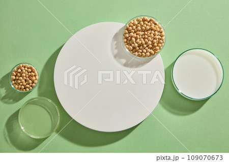 Flat lay of some glass petri dishes of soybean seeds, milk and oil displayed on green background with a round podium. Mock-up for exhibitions or presentation of cosmetic products or packaging Flat lay of some glass petri dishes of soybean seeds, milk and oil displayed on green background with a round podium. Mock-up for exhibitions or presentation of cosmetic products or packaging 109070673