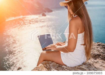 Digital nomad, woman in the hat, a business woman with a laptop sits on the rocks by the sea during sunset, makes a business transaction online from a distance. Freelance, remote work on vacation. 109075003