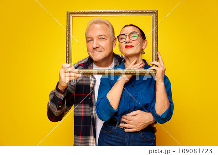 Portrait of happy, loving, senior couple, man and woman posing with picture frame against yellow studio background. Valentine's Day 109081723