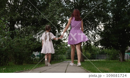 Young mother holds little daughter hand walking on path across green summer park 109081724