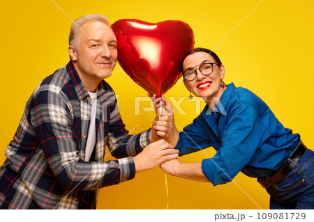 Beautiful, happy, senior couple, man and woman celebrating romantic holiday, holding balloon in heart shape against yellow background. Valentine's Day Beautiful, happy, senior couple, man and woman celebrating romantic holiday, holding balloon in heart shape against yellow background. Valentine's Day 109081729