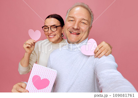 Selfie time. Happy, smiling senior man and woman taking holiday photo with paper heart postcards against pink studio background. Valentine's Day Selfie time. Happy, smiling senior man and woman taking holiday photo with paper heart postcards against pink studio background. Valentine's Day 109081761