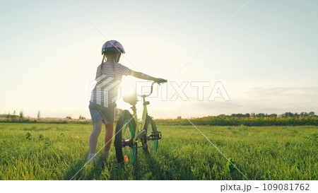 Cute little girl wearing helmet walks pulling bicycle across summer field Cute little girl wearing helmet walks pulling bicycle across summer field 109081762
