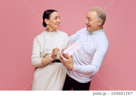 Loving and caring man, husband surprising his beautiful wife with present against pink studio background. Celebration. Valentine's Day Loving and caring man, husband surprising his beautiful wife with present against pink studio background. Celebration. Valentine's Day 109081767