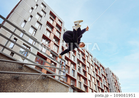 Gymnast, athlete man, young guy jumping, flips of wall, practice Parkour in public park among high-rise buildings over sunrise. 109081951