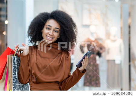 Portrait of a young beautiful woman in a supermarket, a large clothing store, smiling, looking at the camera, holding a phone and colorful packages with shopping gifts. Portrait of a young beautiful woman in a supermarket, a large clothing store, smiling, looking at the camera, holding a phone and colorful packages with shopping gifts. 109084686