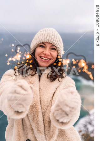 Outdoor winter portrait of happy smiling woman, light faux fur coat holding heart sparkler, posing against sea and snow background 109084688