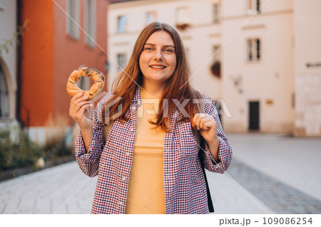 Attractive young female 30s tourist with backpack is holding prezel, traditional polish snack on the Market square in Krakow. Traveling Europe in spring. Selective focus. High quality photo 109086254