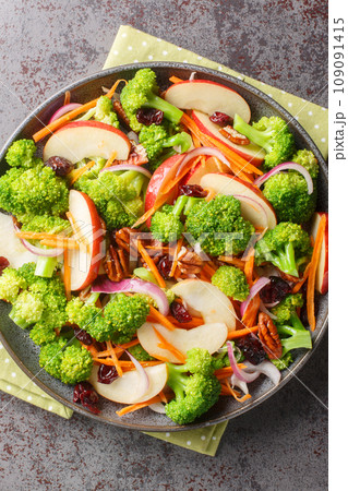 Broccoli apple salad made with fresh broccoli, red apples, carrots, dried cranberries, red onion, nuts closeup on the plate on the table. Vertical top view Broccoli apple salad made with fresh broccoli, red apples, carrots, dried cranberries, red onion, nuts closeup on the plate on the table. Vertical top view 109091415