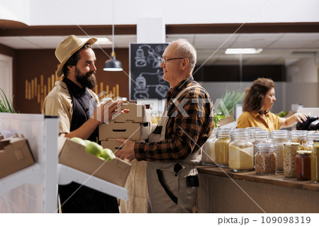 Farmer providing locally grown ecofriendly vegetables to seller selling it to environmentally conscious customers in zero waste shop. Older man handing crates full of vegan produce to retailer Farmer providing locally grown ecofriendly vegetables to seller selling it to environmentally conscious customers in zero waste shop. Older man handing crates full of vegan produce to retailer 109098319
